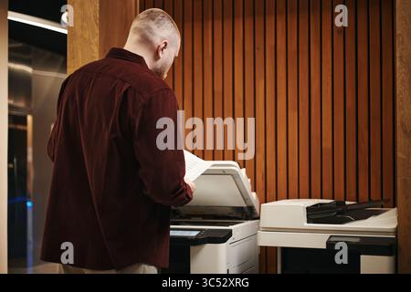 Young man operating office printer standing in modern coworking workspace holding documents preparing to make copies or scan paperwork using multifunction device Stock Photo