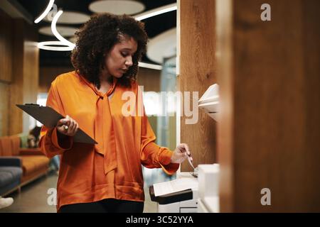 Young Black woman operating photocopier while holding clipboard in modern office setting, standing near wooden partition and focusing on copying documents for work tasks Stock Photo