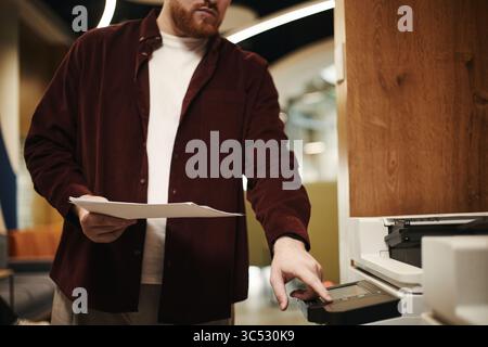 Man operating printer while holding documents in modern coworking office setting, focusing on using device for copying or scanning paperwork, face partially visible Stock Photo