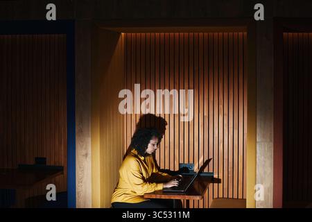 Young Black woman sitting alone using laptop in modern booth workspace, focused on screen, hands on keyboard, wooden slat wall background, side profile visible Stock Photo