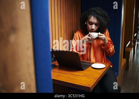 Young Black woman sitting at table using laptop and drinking from cup, focused on screen, working or studying in modern workspace, plate on table, wood panel background Stock Photo