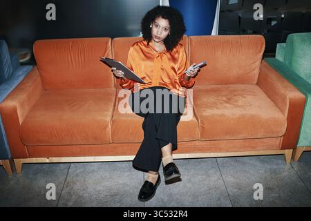 Young Black woman sitting on orange sofa holding tablet and clipboard, looking to side with thoughtful expression, crossing legs, modern office environment visible in background Stock Photo