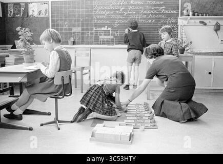 Children in a class preparing something for their teacher Whizgle News, Dutch Desk, The Netherlands, 1950-2000 A classroom scene depicting children engaged in various activities. One child is focused on writing at a desk, while another is kneeling to arrange materials on the floor. A teacher is organizing items nearby, and two other students are standing at a chalkboard covered with writing. The environment appears educational and collaborative, fostering learning and interaction among the students. Stock Photo