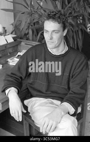 Mr A. de Jong, cultural council, 31-03-1993 Whizgle News, Dutch Desk, The Netherlands, 1950-2000 A man sits at a wooden desk, casually dressed in a dark sweater over a collared shirt, with his hands resting thoughtfully on his knees. Lush greenery is visible in the background, adding a touch of nature to the indoor setting. Stock Photo