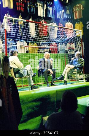 Start Sports Café, Studio, Haarlem, Grote Markt, The Netherlands, 02-01-1998 Whizgle News, Dutch Desk, The Netherlands, 1950-2000 Three individuals are seated in front of a football goal, engaged in a discussion. Behind them, various sports jerseys hang as a backdrop, creating an energetic atmosphere. The focus is on their conversation, with the audience engaged in the scene. Stock Photo