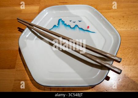 A white hexagonal plate with a blue mountain and bird design and brown chopsticks resting on it, all on a warm wooden surface. Stock Photo
