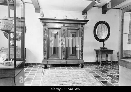 Showroom with antique furniture and pottery, 00-00-1971 Whizgle News, Dutch Desk, The Netherlands, 1950-2000 A spacious interior showcases an intricately carved wooden cabinet as the centerpiece, flanked by display cases holding decorative items. A round mirror hangs on the wall, reflecting the room’s elegant furnishings and tiled floor, creating a harmonious blend of artistry and function. Stock Photo