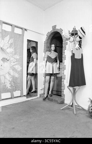 Intr. Fa. Boutiflou, Interior, 13-03-1969 Whizgle News, Dutch Desk, The Netherlands, 1950-2000 A stylish model stands gracefully in a doorway, wearing a chic color-block dress. She leans against the wall, striking a pose, while a mirror reflects her presence. Nearby, a stylish garment rack displays hats and clothing, adding to the fashionable ambiance of the setting. Artwork with intricate designs hangs on the wall, enhancing the creative atmosphere. Stock Photo