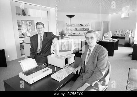 Computer Workshop, H'lem, Haarlem, The Netherlands, 29-11-1991 Whizgle News, Dutch Desk, The Netherlands, 1950-2000 Two men in business attire pose confidently in a modern office space. One leans casually against a computer setup that includes a monitor and keyboard, while the other sits at a desk with a printer nearby. The environment features sleek furnishings and shelves displaying various materials, suggesting a focus on technology and innovation. Stock Photo