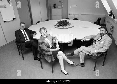 Company Rietveld office furniture, Haarlem, The Netherlands, 30-06-1991 Whizgle News, Dutch Desk, The Netherlands, 1950-2000 A group of three professionals sits around a distinctive octagonal conference table in an office setting, exuding a collaborative atmosphere. The individuals are dressed in formal business attire, engaging in a moment of discussion or reflection within a well-organized workspace. A small plant adorns the center of the table, adding a touch of warmth to the environment. Stock Photo