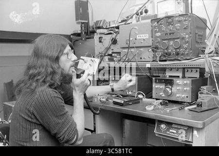 Radio amateurs IJmuiden, Radio, 15-07-1978 Whizgle News, Dutch Desk, The Netherlands, 1950-2000 A person with long hair is engaged in radio communication, speaking into a handheld device while adjusting dials and buttons on an array of vintage radio equipment stacked on a cluttered workbench. Various electronic components and tools are also present, illustrating a focus on technology and amateur radio operation. Stock Photo