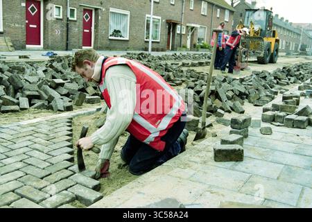 Student road builders project, IJmuiden, IJmuiden, The Netherlands, 20-03-2000 Whizgle News, Dutch Desk, The Netherlands, 1950-2000 A construction worker kneels on a partially paved street, carefully laying bricks while others in the background operate machinery and prepare additional stones. The scene captures the effort and teamwork involved in urban roadwork. Stock Photo