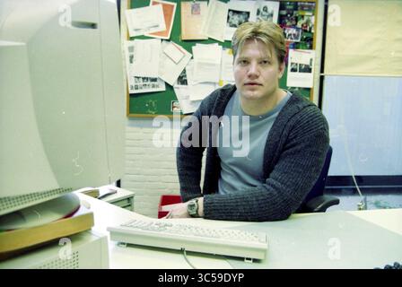 Dennis van der Geest. for column, Haarlem, among others, Haarlem, The Netherlands, 23-02-2000 Whizgle News, Dutch Desk, The Netherlands, 1950-2000 A young man with blonde hair sits at a desk wearing a gray shirt and a dark cardigan. He appears to be focused while looking directly at the viewer. Behind him, a bulletin board is filled with papers and notes, indicating an organized workspace. A computer monitor and keyboard are positioned in front of him. Stock Photo