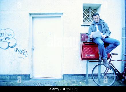 Pim Bijtel, Haarlem, The Netherlands, 06-03-2001 Whizgle News, Dutch Desk, The Netherlands, 1950-2000 A man casually sits atop a red mailbox, holding a stack of papers as he leans against a blank wall. Nearby, a bicycle rests on the ground, adding to the relaxed urban scene, while graffiti decorates the background. Stock Photo