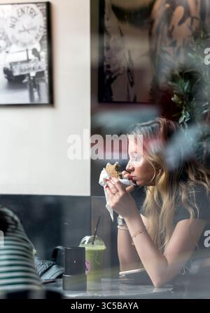 Young woman eating in a London cafe, seen through the window, with reflections and soft focus—urban lifestyle and candid street photography. Stock Photo