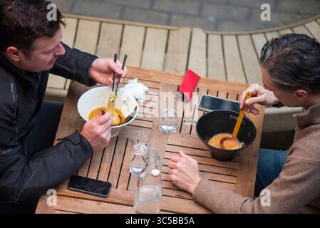 Two men eating ramen noodles outdoors at a wooden table in Soho, London, using chopsticks and soup spoons. Stock Photo