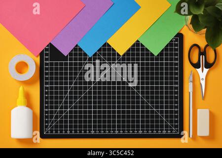 A black cutting mat with white grid lines sits on a bright yellow background surrounded by neatly laid out art hobby supplies: pencil, glue, tape, sci Stock Photo