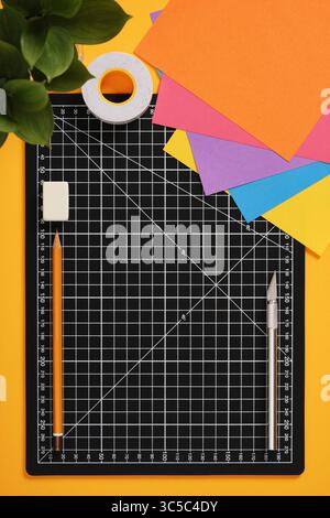 A black gridded cutting mat on a bright yellow surface, surrounded by colorful paper sheets, a precision knife, white tape, an orange pencil, and a re Stock Photo