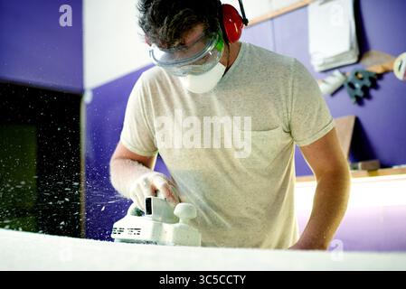 Design, sanding and surfboard with man in workshop for manufacturing or production process. Earmuffs, face mask and goggles with person shaping Stock Photo