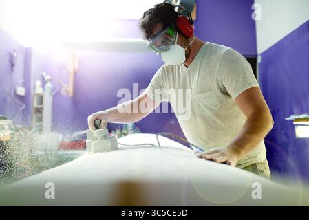 Man, tool and sanding surfboard in workshop for grinding process, shape surface and custom design. Male person, equipment and prepare for product Stock Photo
