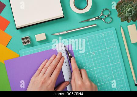 Hands working with colorful paper using a paper knife on mint green cutting mat. Tools for paper crafts. Ruler, pencil, scissors, tape, eraser Stock Photo