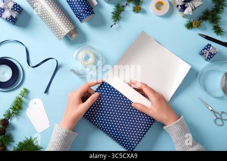 Hands wrap the box in wrapping paper. Around it are ribbon, scissors, tape, spruce twigs, and small wrapped gifts. Preparing for the holidays. Stock Photo