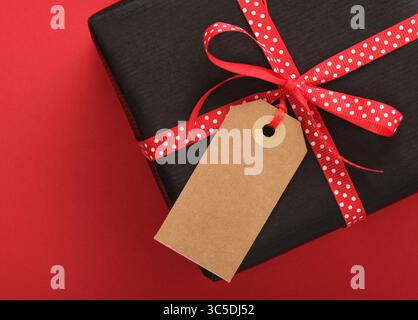 Card with black craft gift box with empty rectangular tag on red background. Place for your text Stock Photo