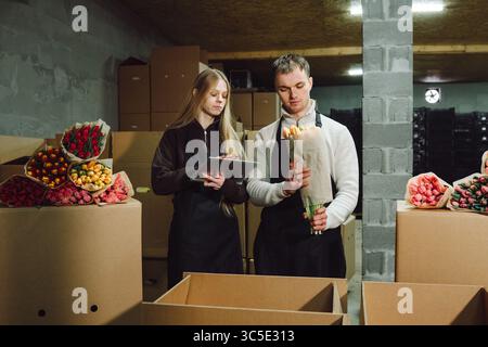 Two floral workers in a warehouse preparing fresh bouquets, surrounded by colorful flowers and cardboard boxes, showcasing teamwork and dedication in Stock Photo