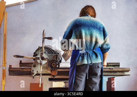 Back view of a young woman wearing a apron and wool sweater operating a vintage printing press  in art classroom studio. Stock Photo