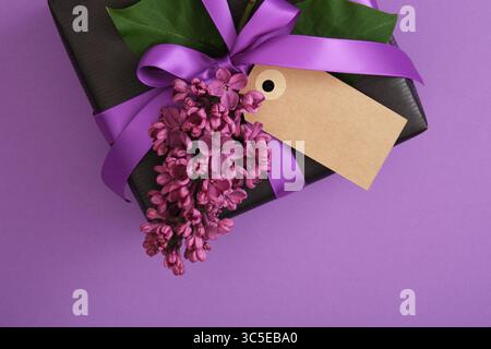 Black craft gift box with empty tag, lilac flowers on violet background. Place for your text Stock Photo