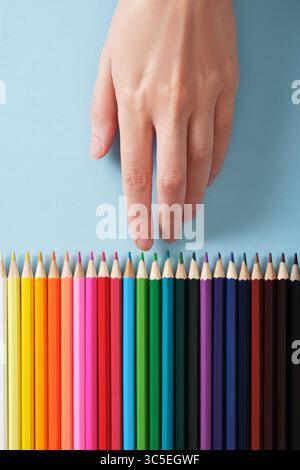 Hand choosing green colored pencil from a row of multi-colored pencils on light blue background. Stock Photo