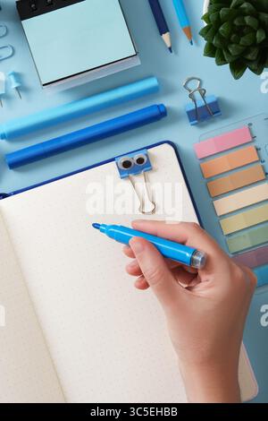 Top view of hand holds a blue felt-tip pen in front of an open notebook with a blank page. Around blue office supplies pens, pencils, felt-tip pens, s Stock Photo