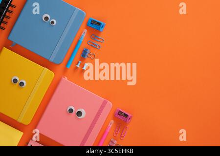 Blue, yellow and pink notebooks with googly eyes and office supplies on bright orange background. Place for your text. Stock Photo