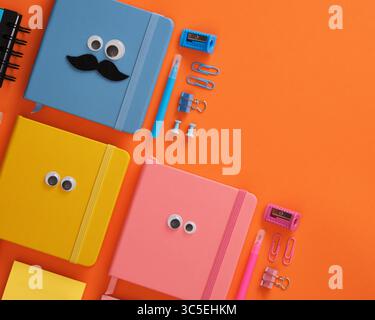 Colorful notebooks with googly eyes and funny office supplies on orange background. Copyspace. Stock Photo