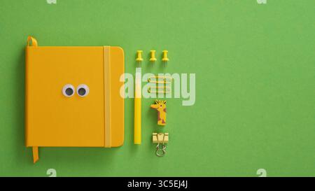 Yellow notepad with eyes and stationery items felt-tip pen, pins, eraser, paper clips on a green background. Space for your text. Stock Photo