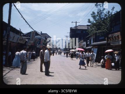 A bustling street scene in Tague (Daegu), South Korea, June 1957. Pedestrians, including men in military uniforms and women in traditional hanbok and mid-century Western attire, walk along a lively commercial street lined with storefront signs and umbrellas under bright summer sunlight. Stock Photo