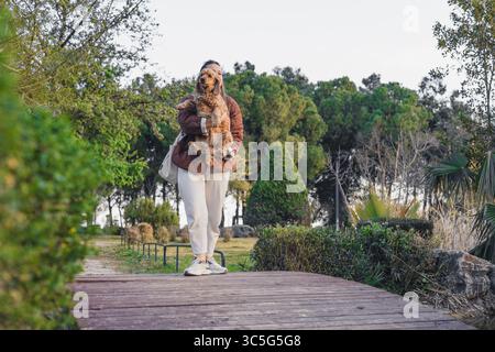 A young woman with long brown hair carries a cocker spaniel dog in a park. The scene features lush greenery and a wooden pathway. Stock Photo