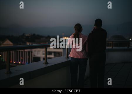 Two people standing on a rooftop balcony overlooking the cityscape at dusk, sharing a quiet reflective moment in an urban environment. Stock Photo