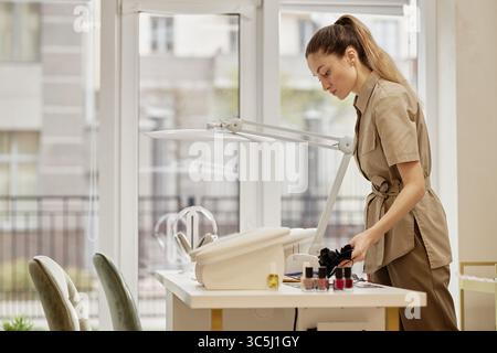 Young adult Caucasian woman preparing manicure tools at nail salon workstation, arranging equipment and nail polish bottles on table with large window Stock Photo