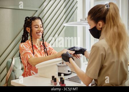 Young adult biracial woman smiling while receiving manicure from young adult Caucasian woman wearing gloves and face mask in nail salon, hands resting Stock Photo