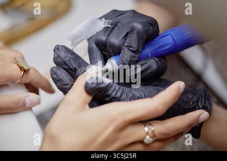 Caucasian young adult woman receiving manicure from gloved hands of young adult woman using electric nail drill, close up on hands during nail care pr Stock Photo