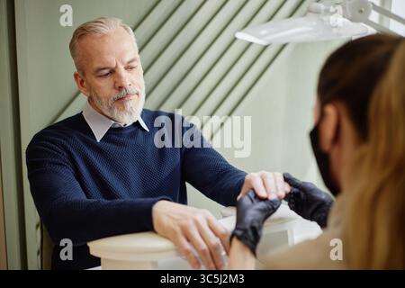 Middle aged Caucasian man receiving manicure from young adult Caucasian woman wearing gloves, man sitting at nail salon table looking at technician, h Stock Photo