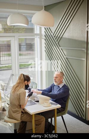 Caucasian senior man receiving manicure from young Caucasian woman in salon, both sitting at table near large window, man holding hands out while woma Stock Photo