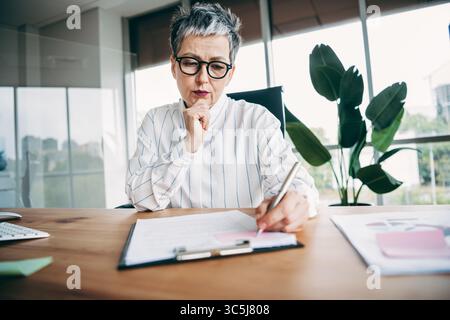 Confident mature businesswoman working on professional documents at her elegant modern office desk during a workday Stock Photo