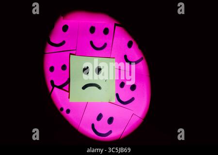 Sad face surrounded by happy faces drawn on colored paper with black marker with a uniform solid background Stock Photo