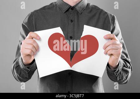 Betrayal. Man tearing sheet of paper with red heart into halves on grey background, closeup Stock Photo