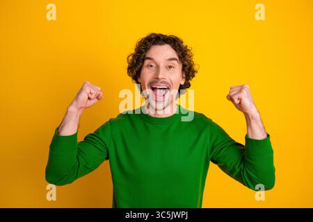Excited young man in a vibrant green sweater celebrating with joyful gestures against a yellow background Stock Photo