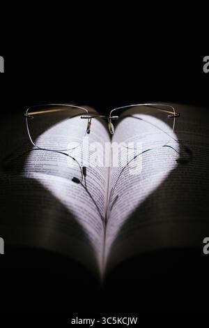 Open book illuminated with heart-shaped light and glasses on a black background. Stock Photo