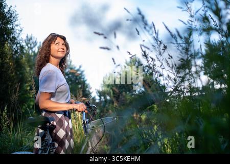 Curly-haired woman stands with bicycle in vibrant green field, surrounded by tall grass and trees, embracing the peacefulness of nature and personal w Stock Photo