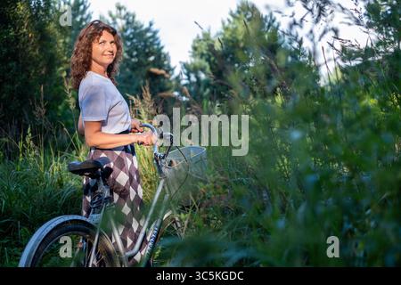Curly-haired woman stands next to bicycle in vibrant greenery, embracing wellness and nature, radiating joy and tranquility in a peaceful outdoor envi Stock Photo